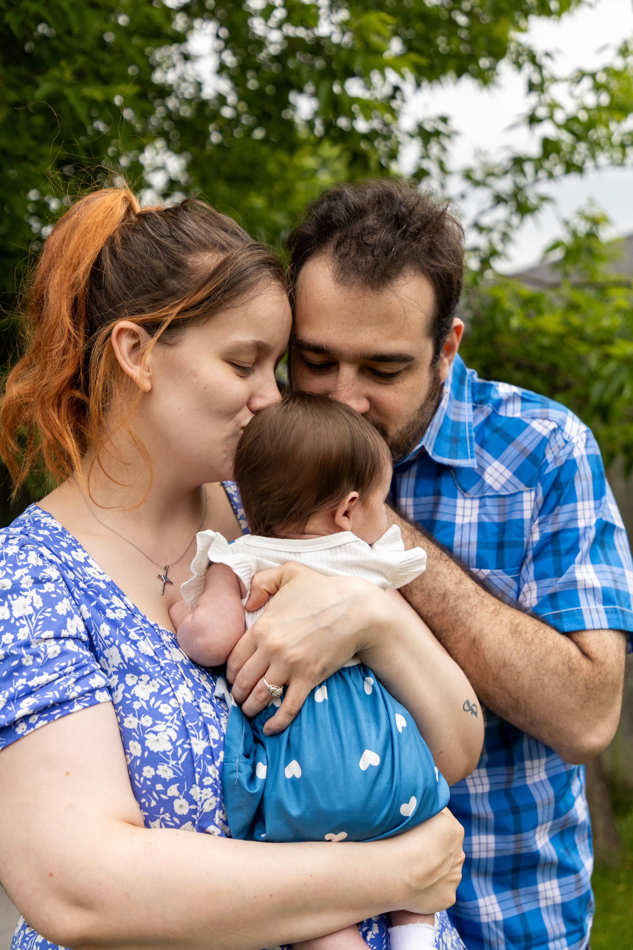 A family photo with 2 new parents. The baby's facing away from the camera and the parents kissing her head