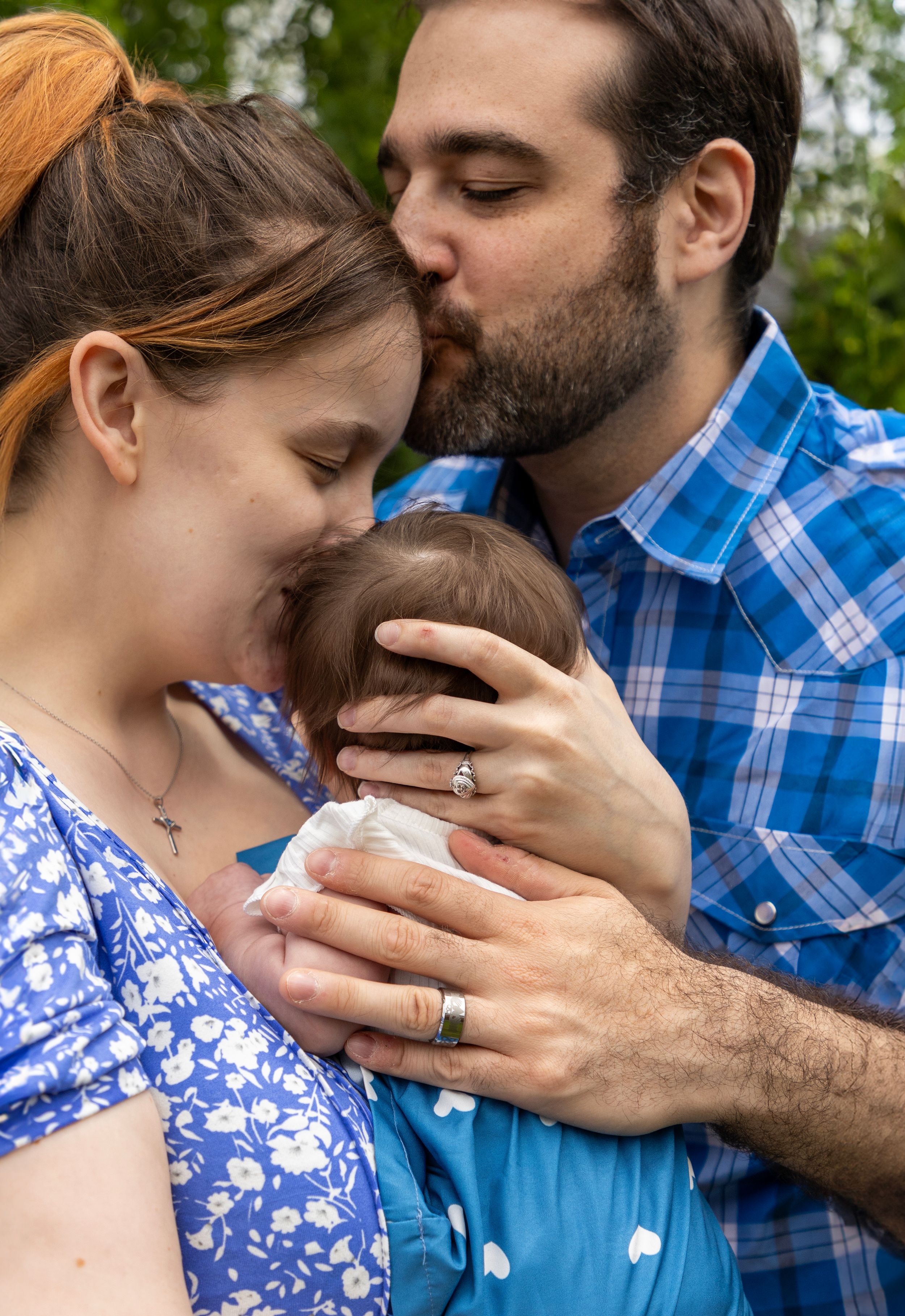 A close up family photo with 2 new parents. The baby's facing away from the camera with mom kissing her head and dad kissing moms forehead