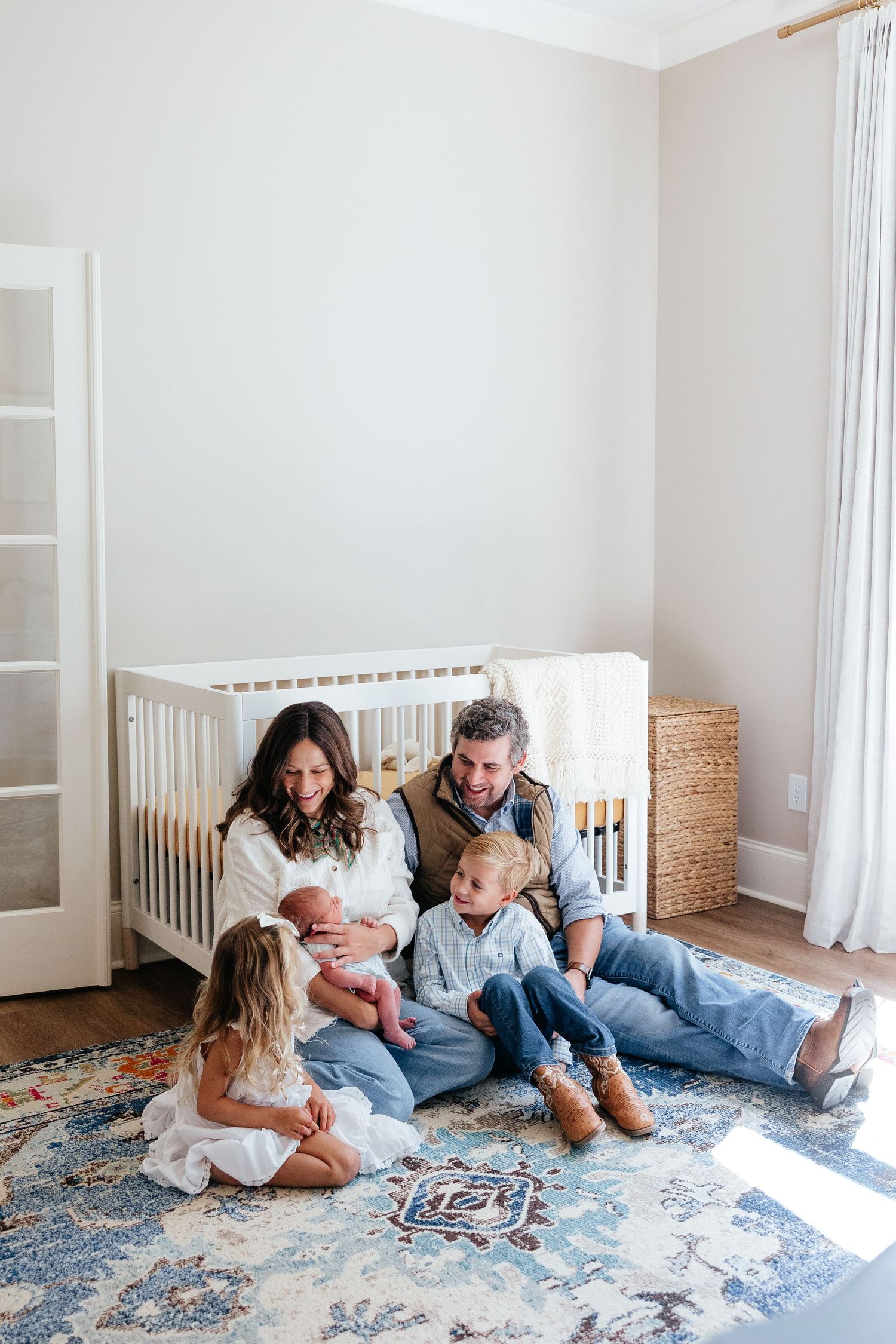 A family relaxes together on a blue patterned rug in a bright, minimalist nursery with white walls and natural light.