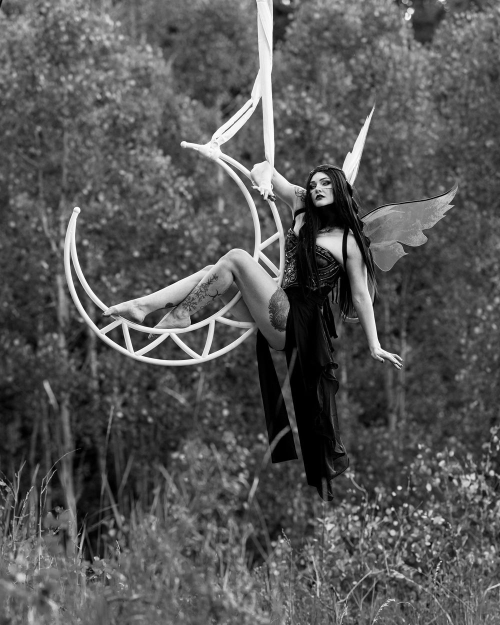 Aerialist in fairy wings on a moon lyra. She is sporting a dark esthetic. Aerial hoop, circus artist, fae, aerial, aerial dance