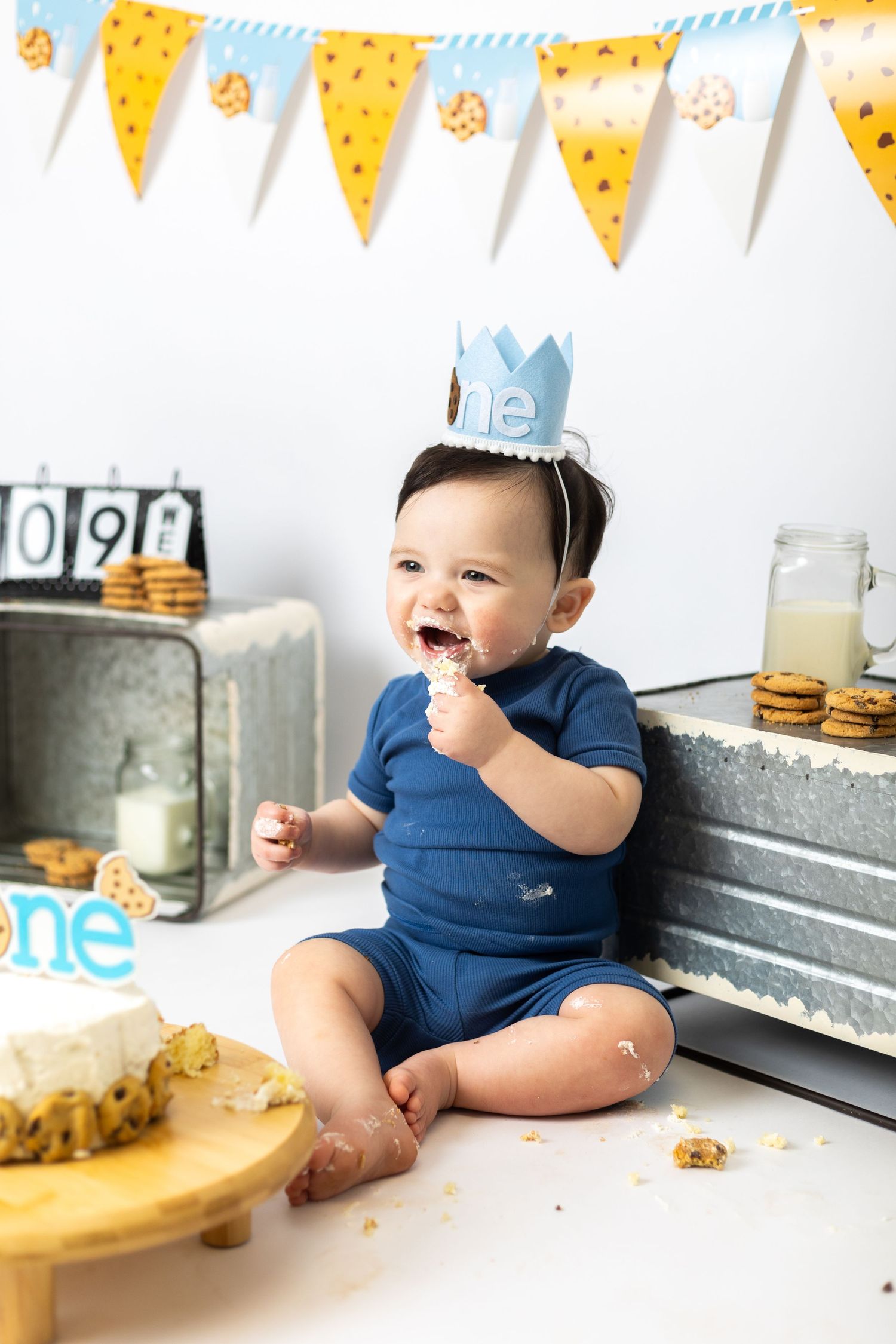 Baby in navy blue outfit sits near cookies and party decorations during first birthday celebration photoshoot.