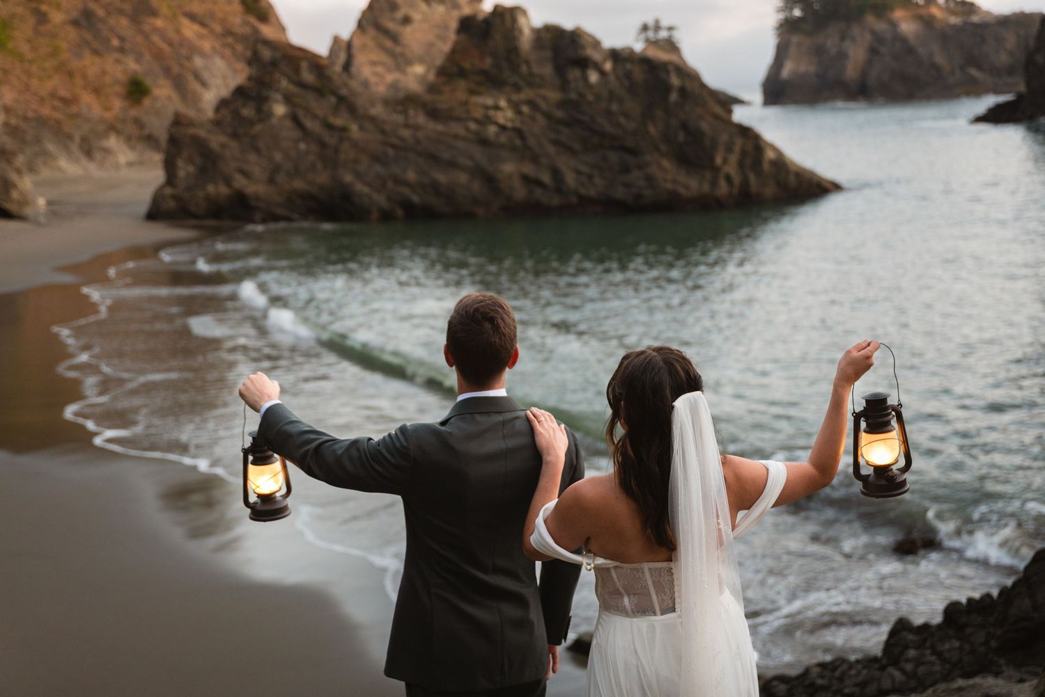 Newlyweds holding vintage lanterns overlooking a rocky coastal beach at dusk.