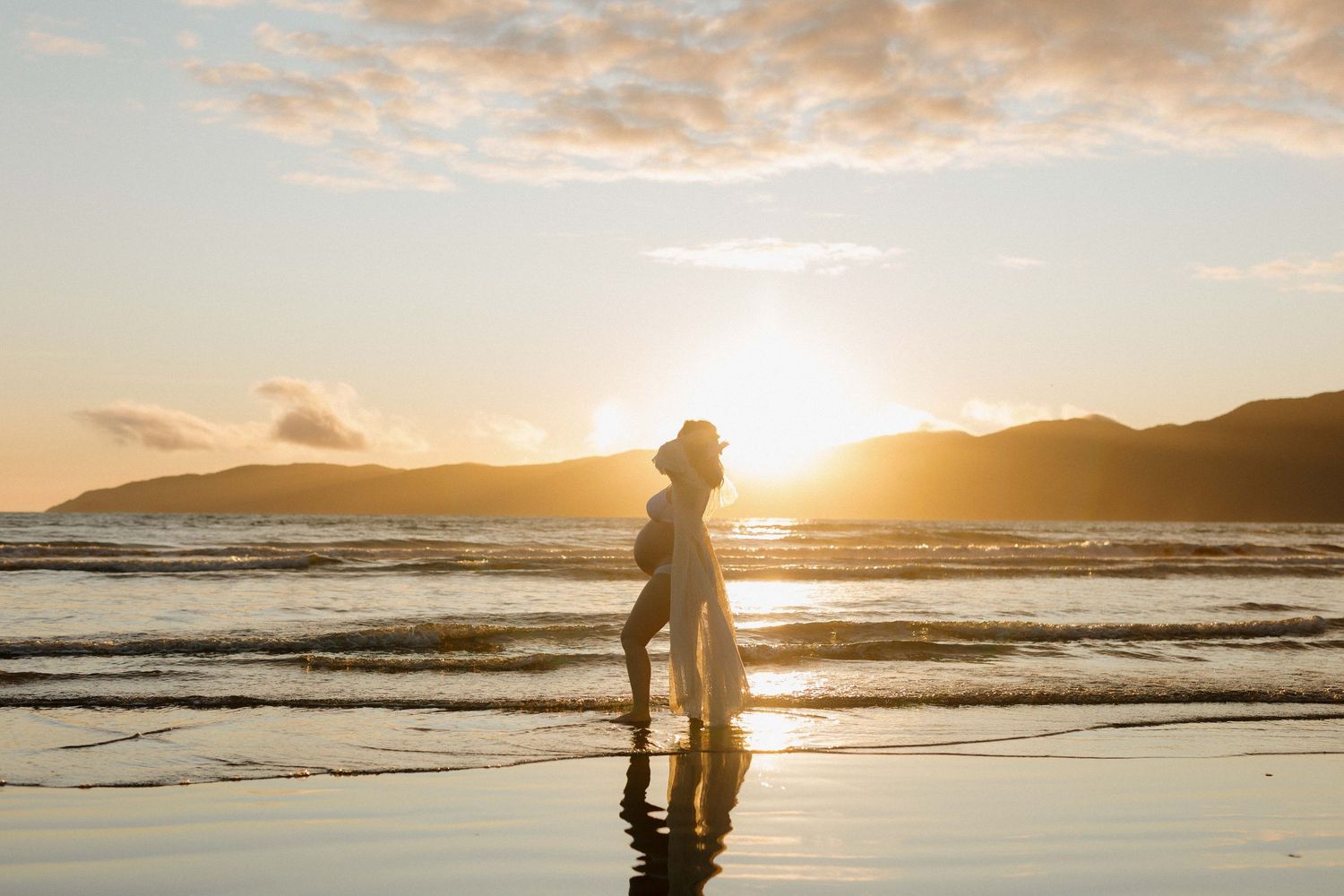 A silhouette stands on a beach at sunset with waves rolling in and mountains in the background.
