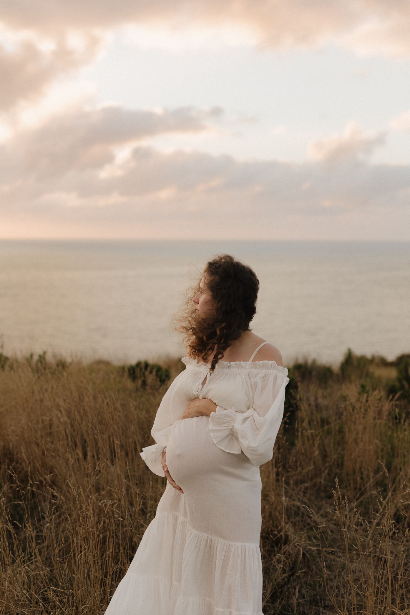 A woman in a flowing white dress stands in golden grass at sunset overlooking the ocean.
