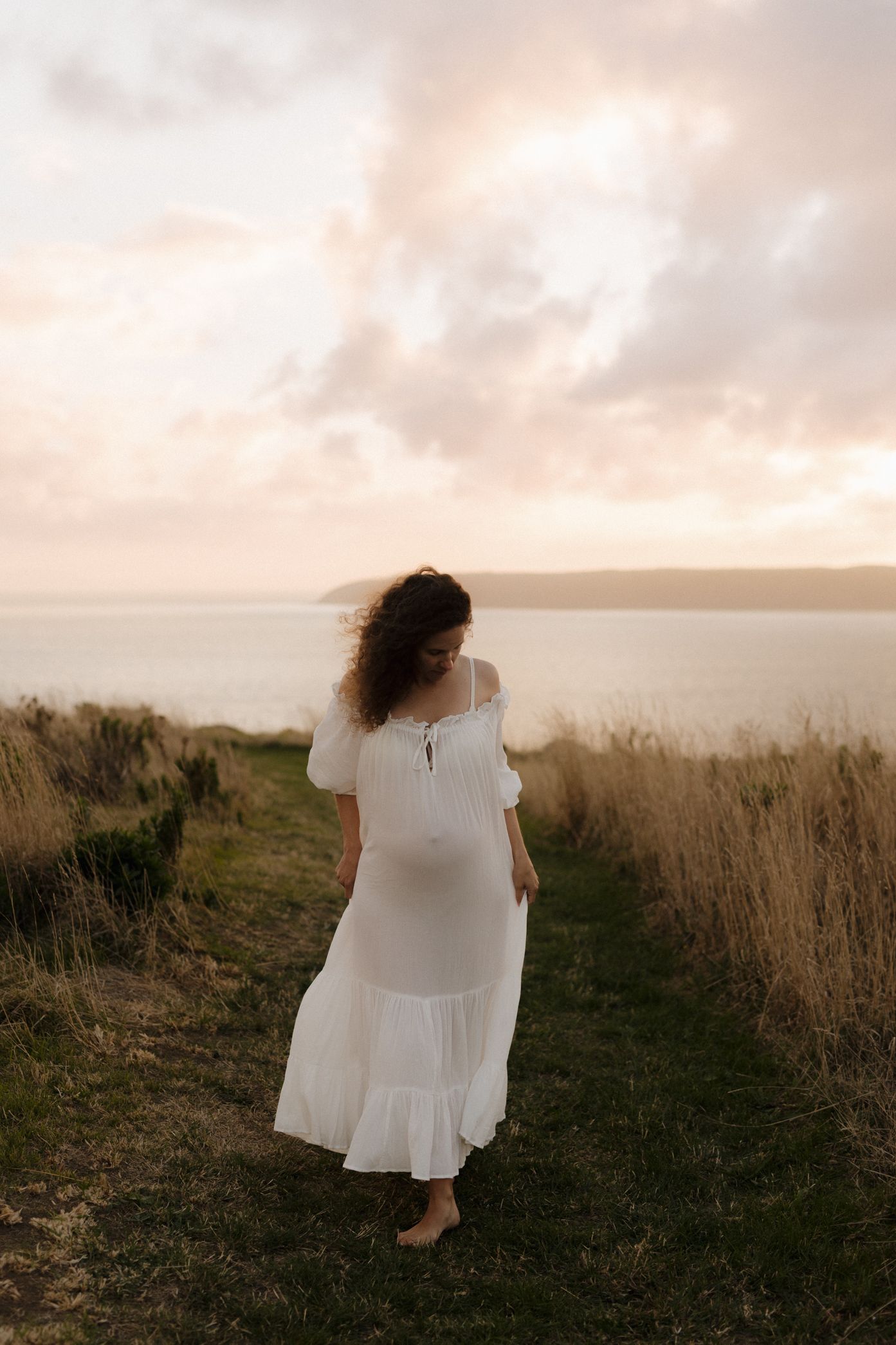 A person in a flowing white dress walks along a grassy coastal path at sunset.