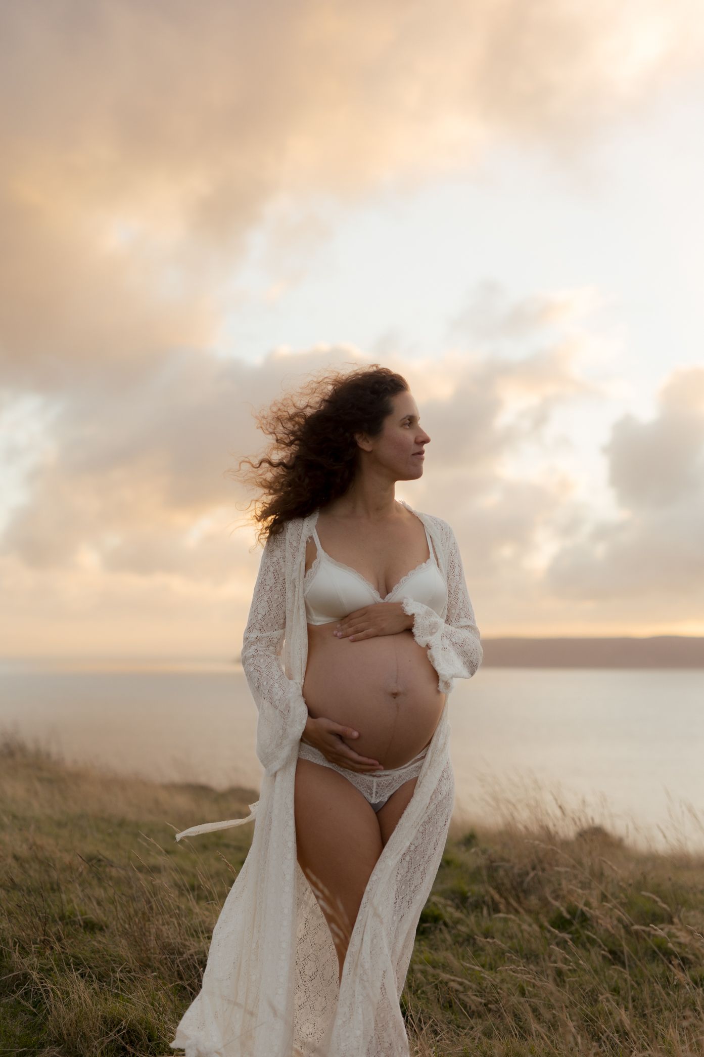 Ethereal maternity photo of an expectant mother in white lace robe standing on coastal cliffs at sunset.