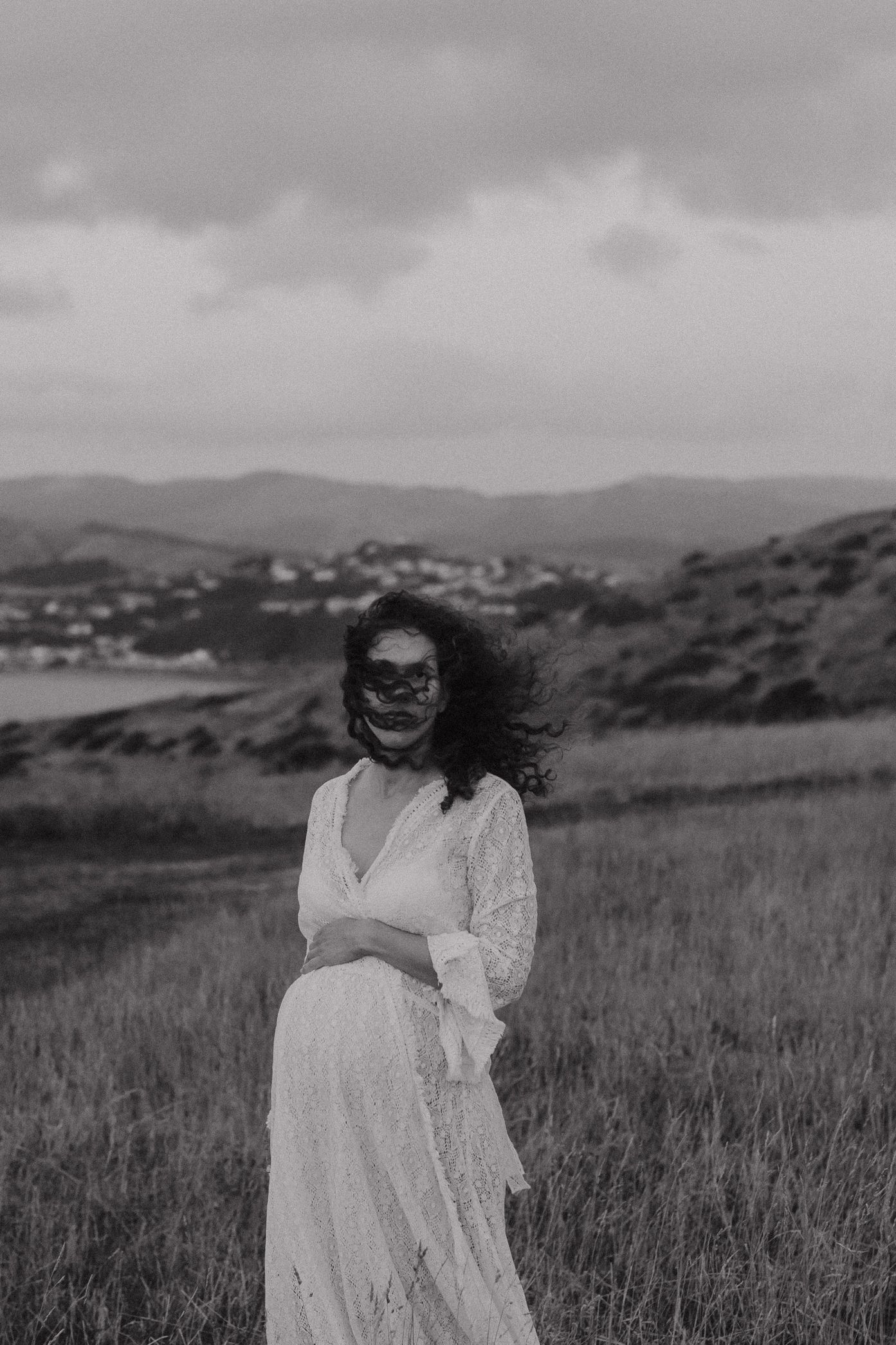 A woman in a white lace dress poses in a grassy field with rolling hills and cloudy skies in the background.