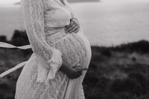 A black and white photograph shows a pregnant figure in a lace dress gently cradling their belly outdoors.