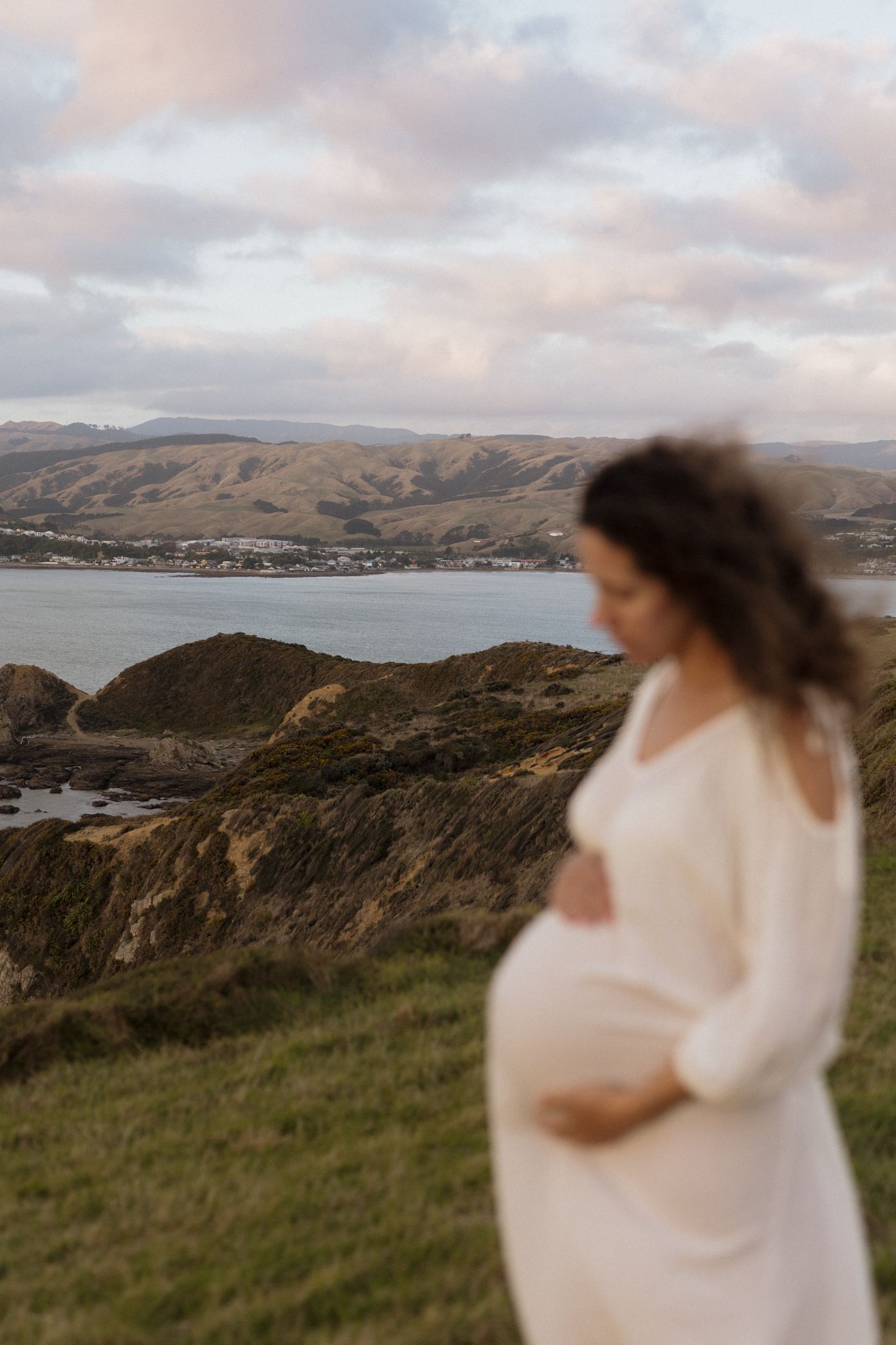 A figure in a white dress stands on a coastal cliff overlooking the ocean and rolling hills at sunset.