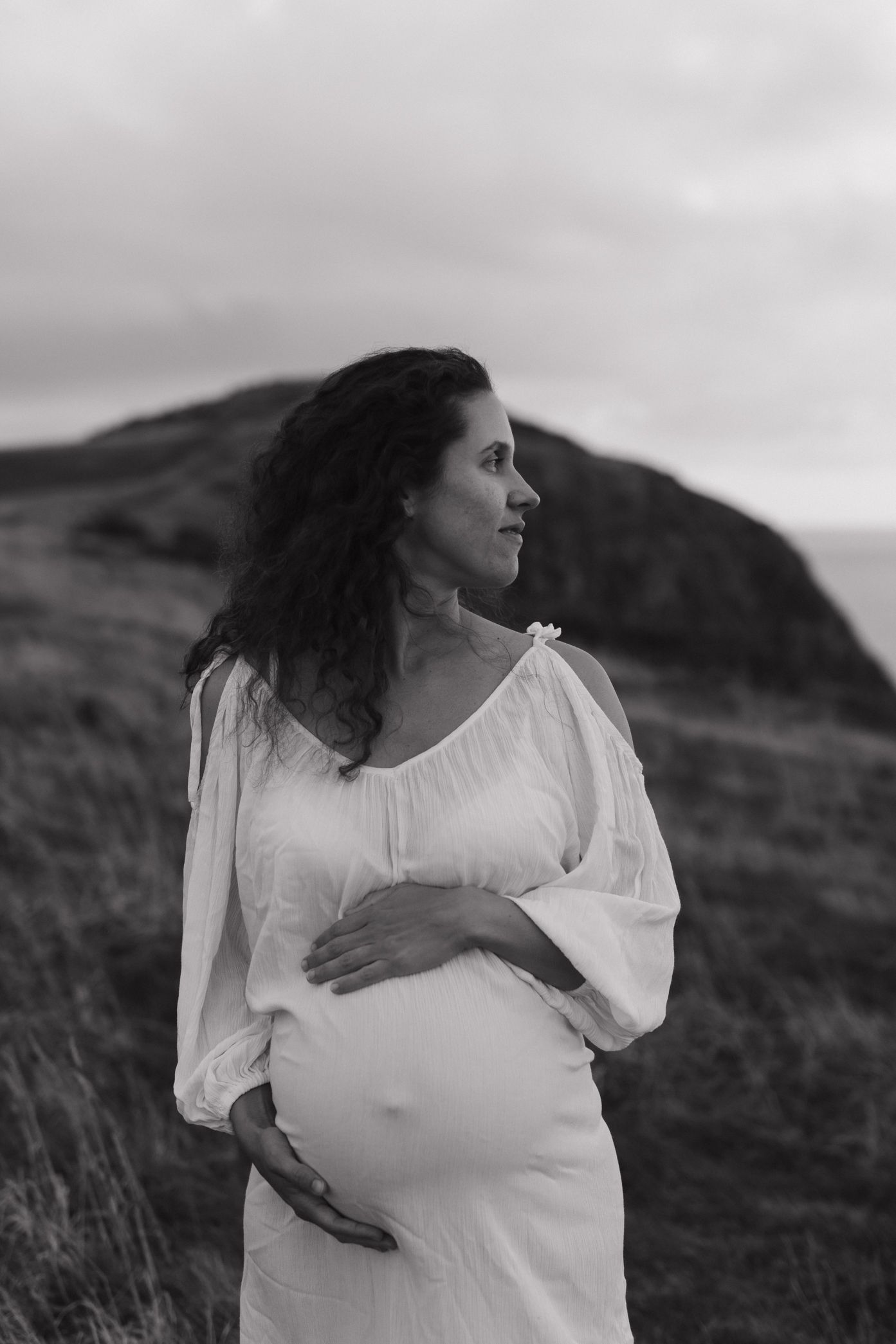 A series of black and white photos showing a pregnant woman in a white dress standing against a rugged hillside landscape.