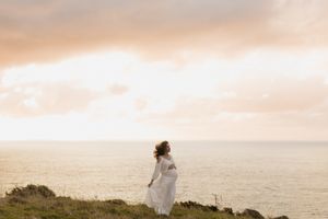 A bride in a flowing white dress stands on a scenic coastal cliff overlooking the ocean at sunset.