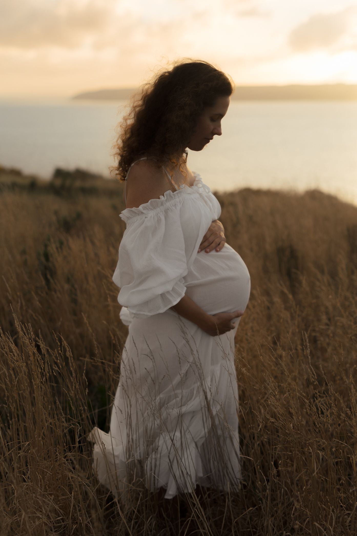 Silhouette of pregnant woman in white dress standing in golden field at sunset.