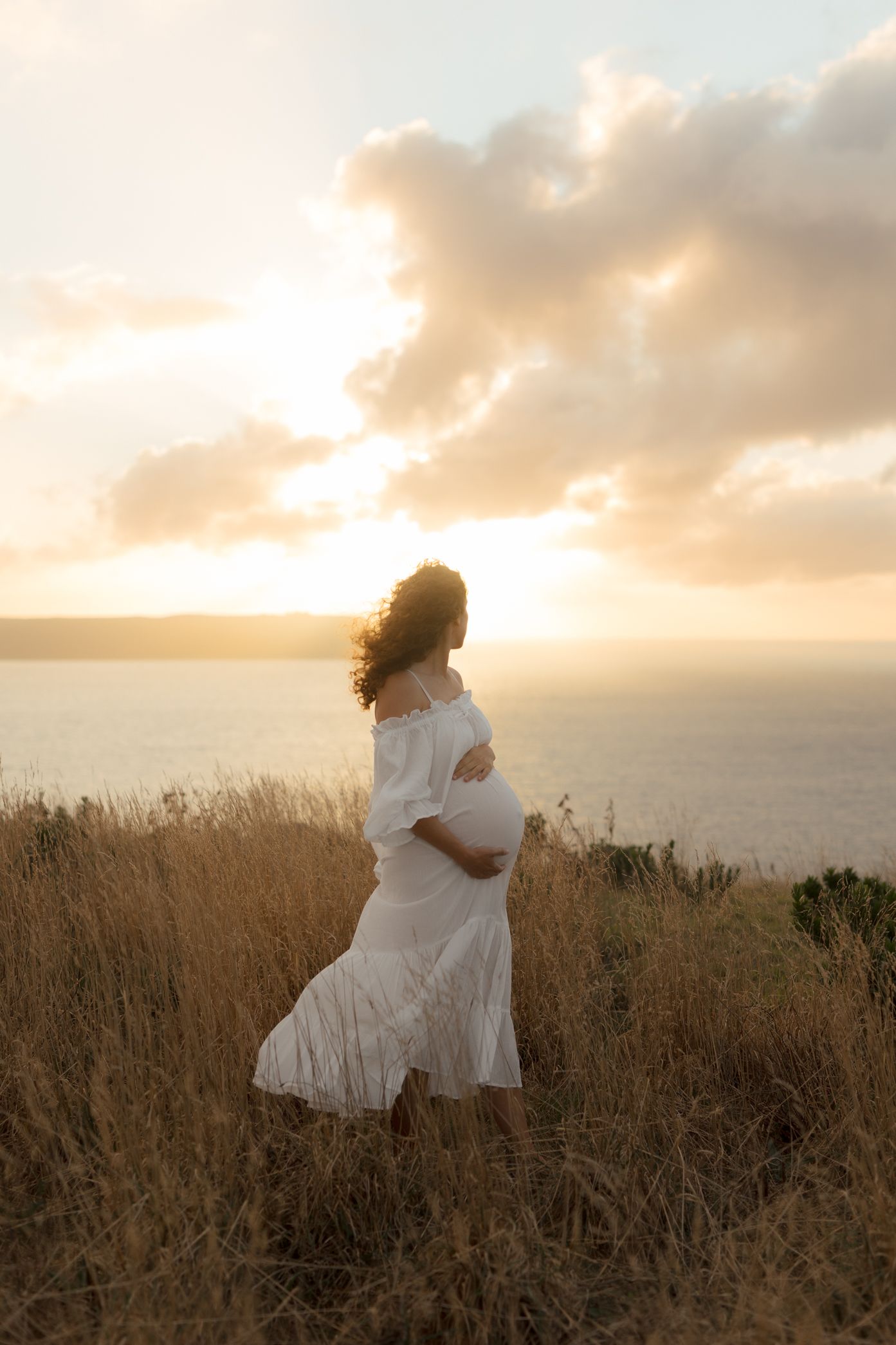 Romantic couple in white clothing embraces against a dramatic sunset sky over a coastal meadow.