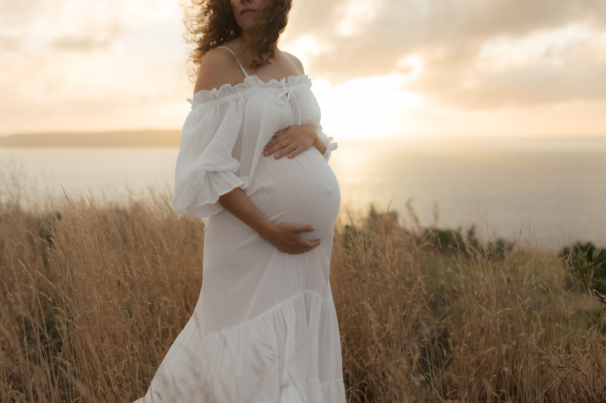 A person in a flowing white dress stands in a golden field at sunset holding their pregnant belly.