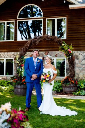 Wedding party poses on green lawn in front of wooden lodge with stone accents and colorful floral decor.