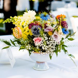 Small floral centerpieces with yellow, pink and blue blooms arranged in white vases on a light table setting.