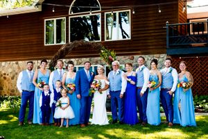 Wedding party in blue dresses and suits poses outdoors against rustic wooden wall backdrop during summer celebration.