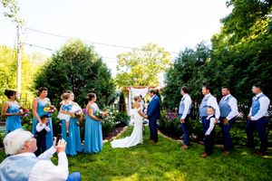 A panoramic wedding ceremony scene in a lush garden setting with guests seated in white chairs at sunset.
