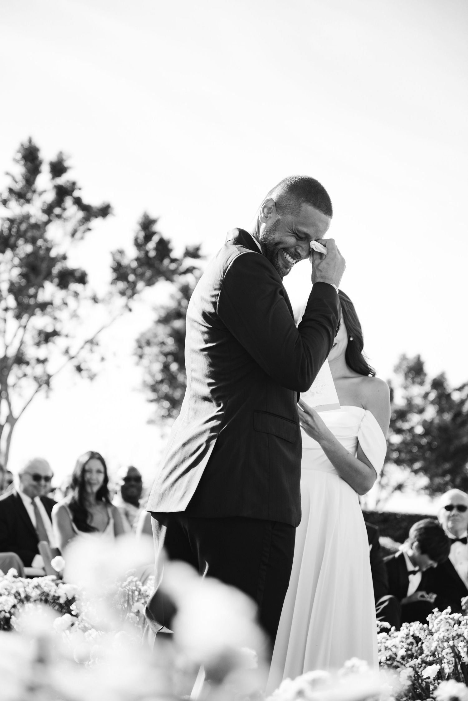 Black and white wedding ceremony photos showing couple exchanging vows outdoors among flowers and guests.