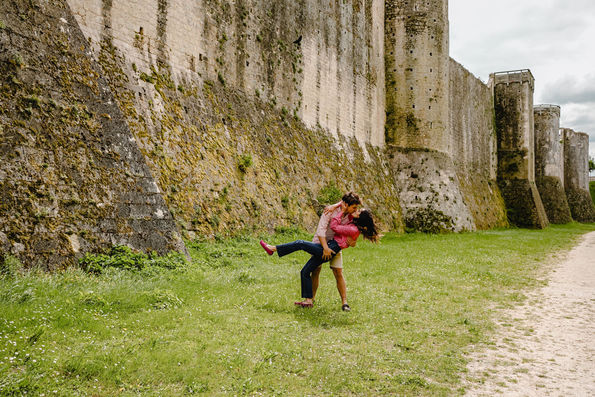 A couple playfully interacts along a stone wall path on a cloudy day.