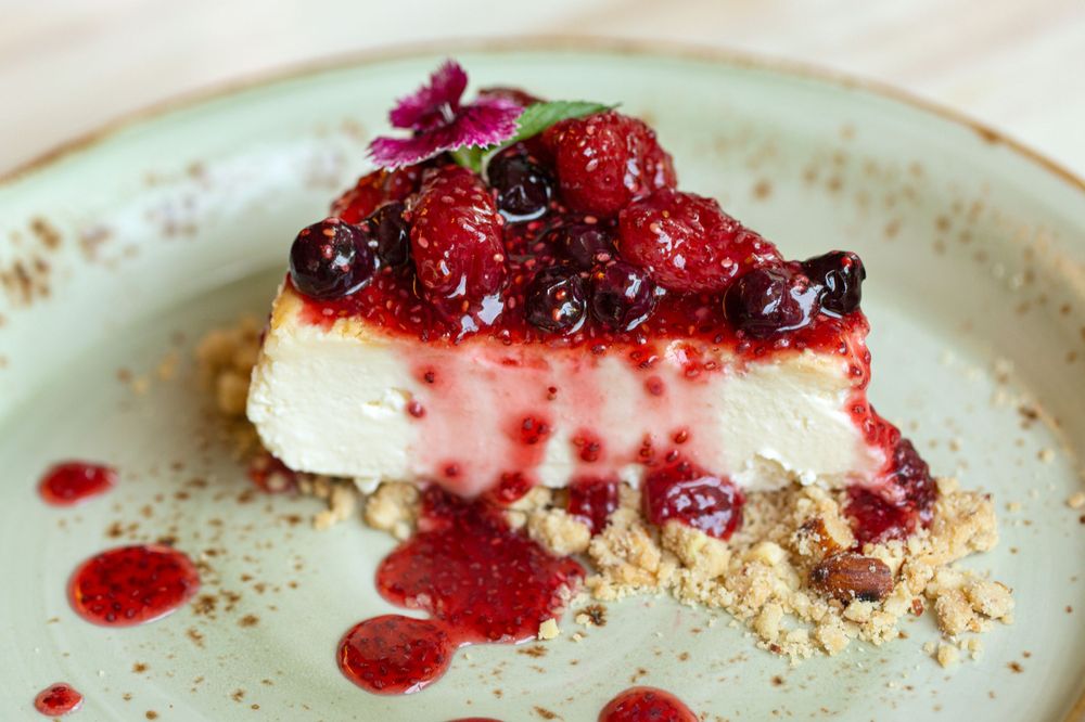 A slice of creamy cheesecake topped with mixed fresh berries and crumble on a ceramic plate with berry sauce.