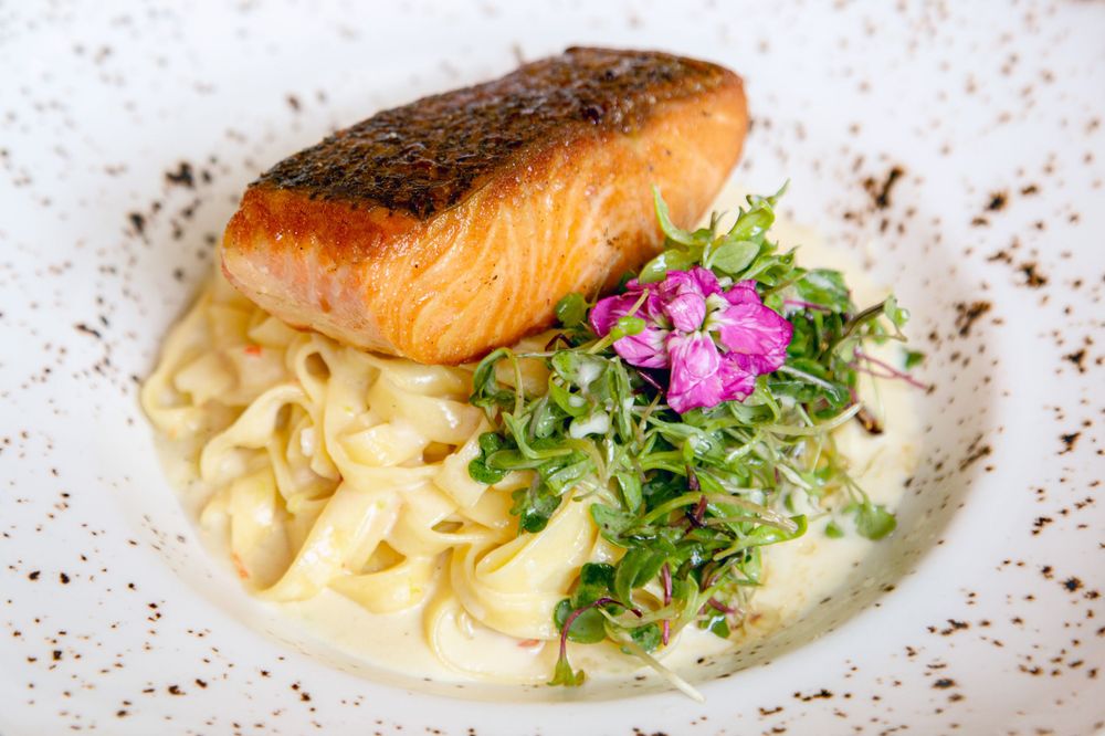 Pan-seared salmon fillet served over creamy fettuccine pasta with microgreens and purple flower garnish on white plate.