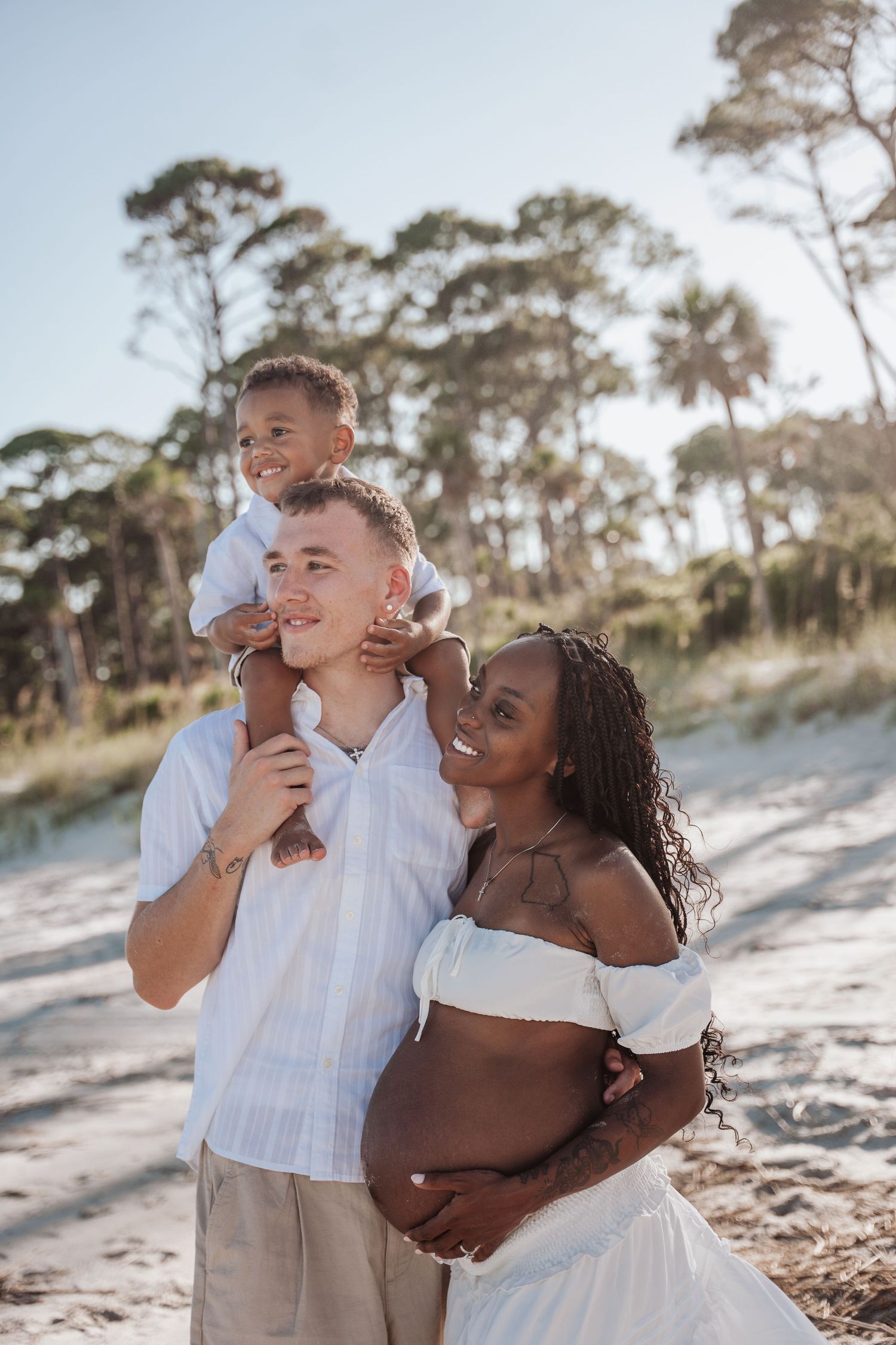 Joyful couple embraces and laughs together during sunset photoshoot on sandy beach with pine trees in background.