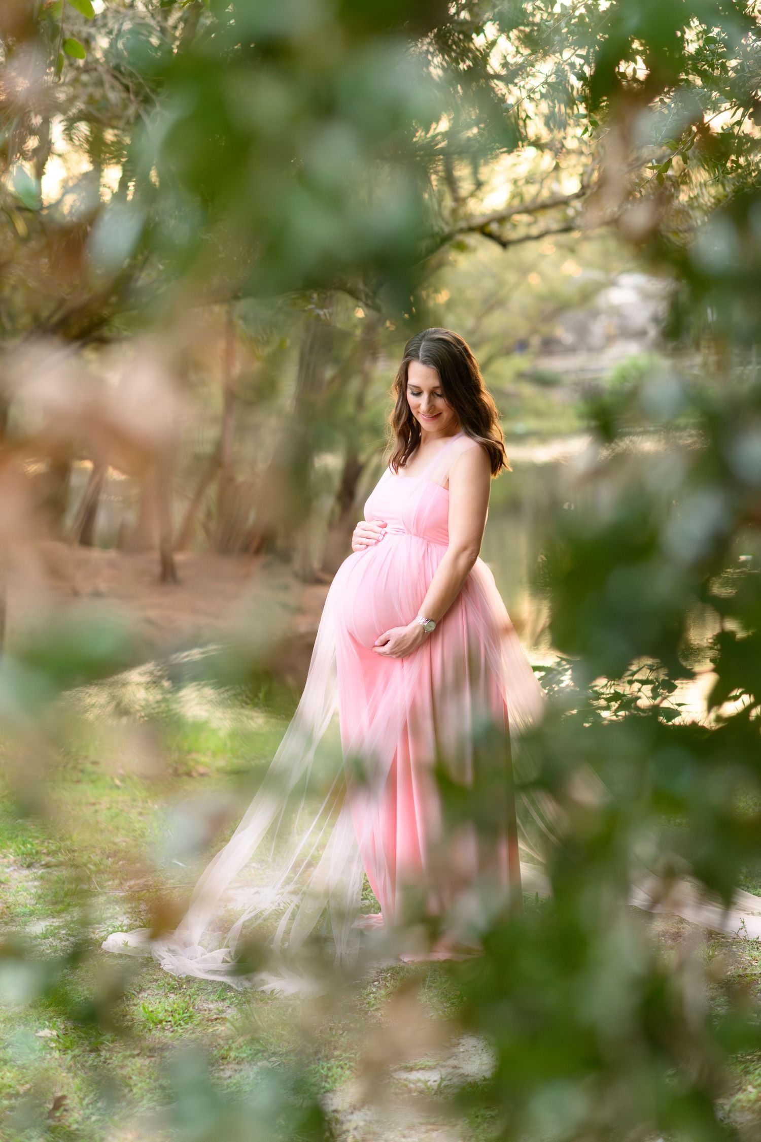 A dreamy maternity photo in a pink flowing dress captured through soft bokeh forest foliage at golden hour.