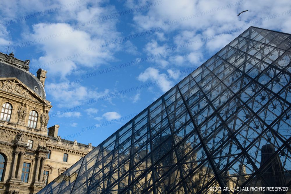 The iconic glass pyramid entrance of the Louvre Museum stands against a blue sky with puffy white clouds in Paris.