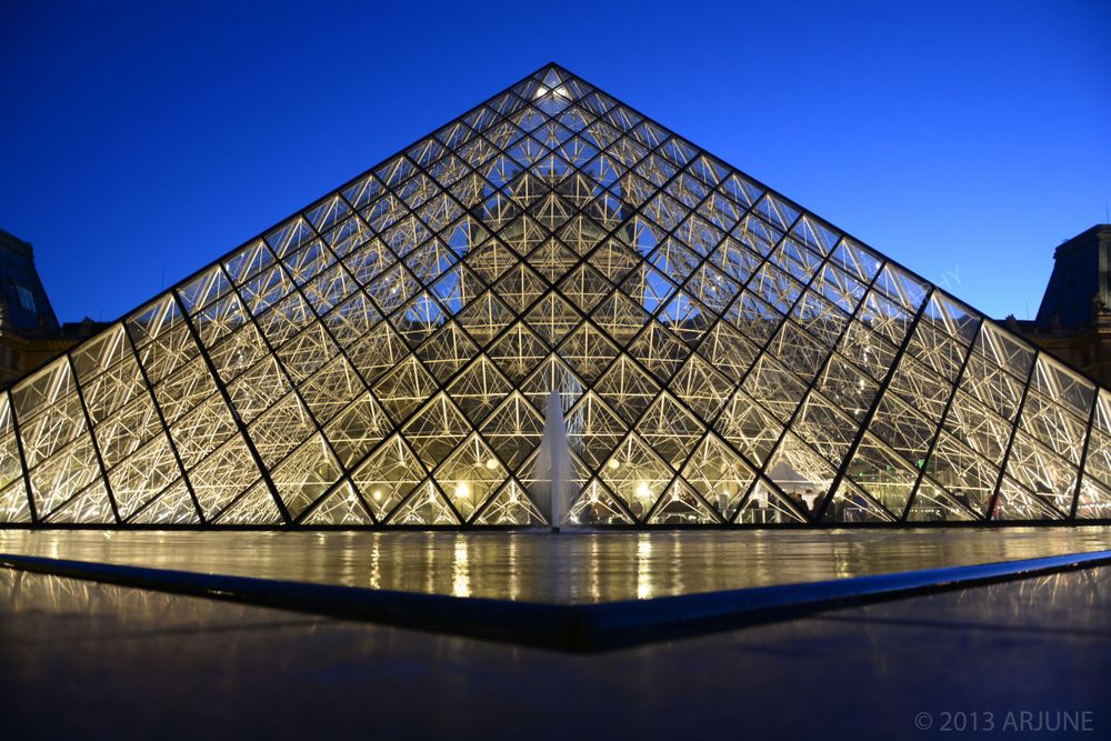 The illuminated glass pyramid entrance of the Louvre Museum in Paris glows against a deep blue twilight sky.