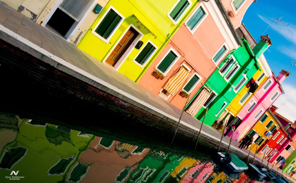 Colorful houses in vibrant yellow, orange, green and pink reflect in the canal waters of Burano, Venice.