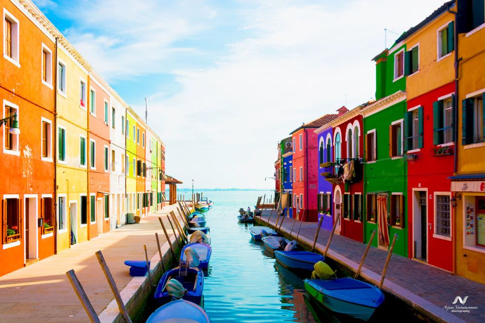 Colorful houses line a canal with moored boats in Burano, Venice, with vibrant orange, pink and green buildings.