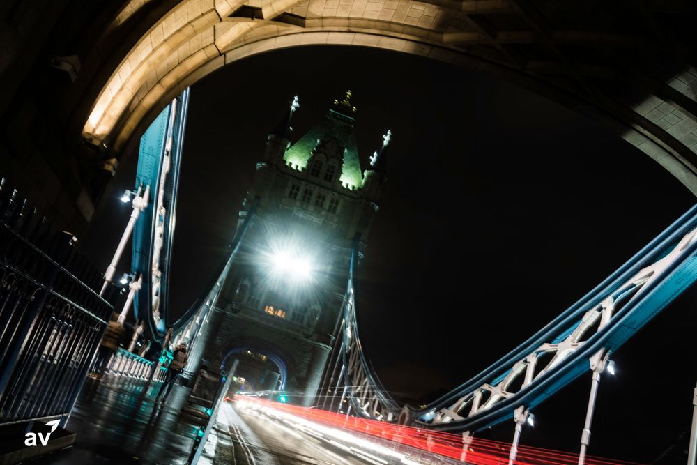 Long exposure night shot of London's Tower Bridge with light trails from traffic and illuminated towers against dark sky.