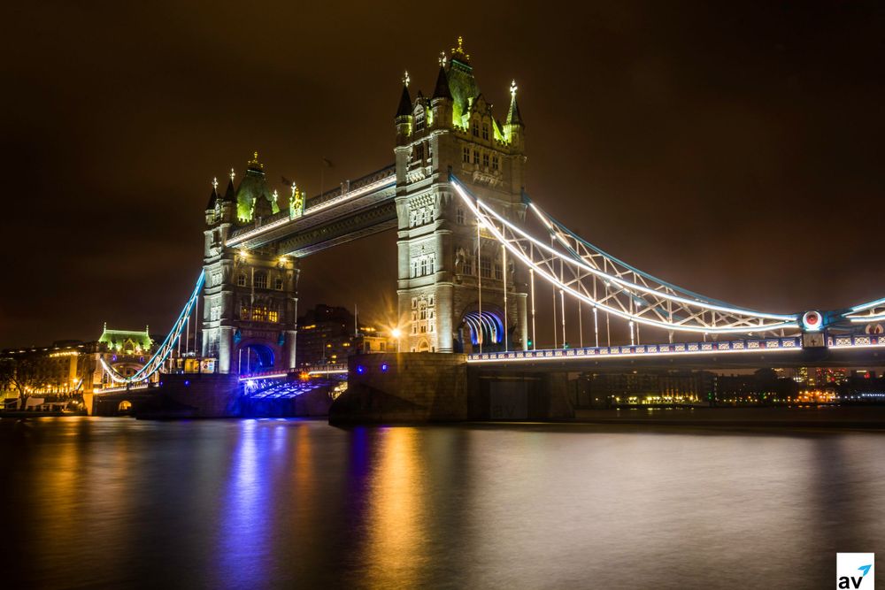 Tower Bridge illuminated at night with blue and green lights reflecting on the River Thames in London.