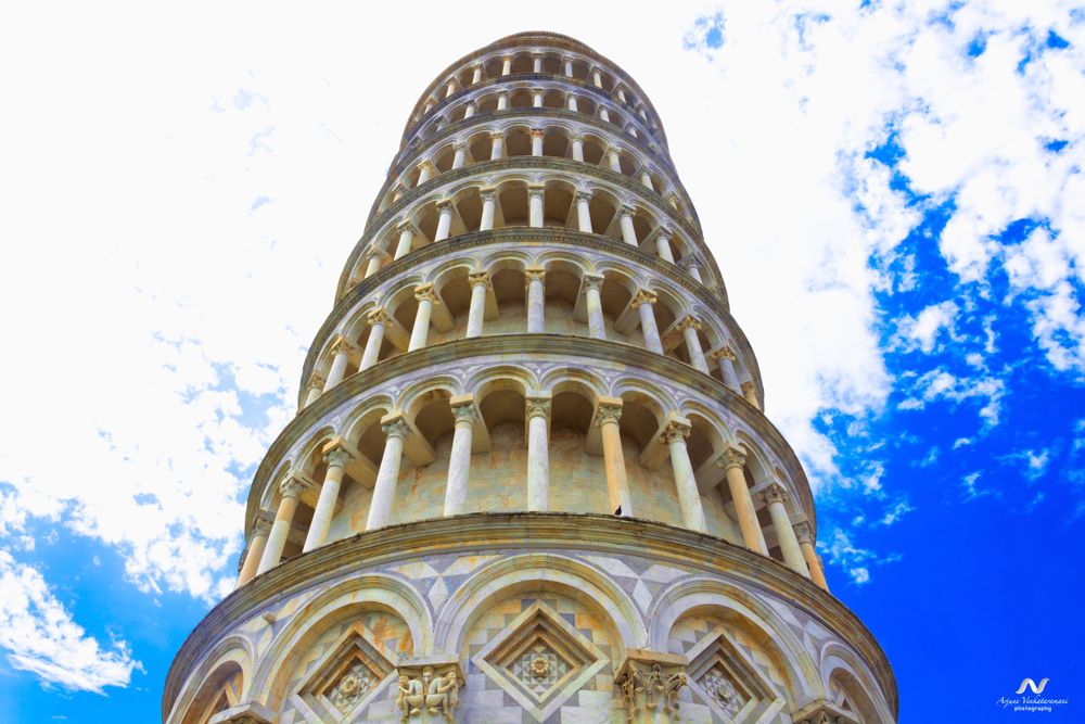 Low angle view of the famous Leaning Tower of Pisa against a bright blue sky with white clouds in Italy.
