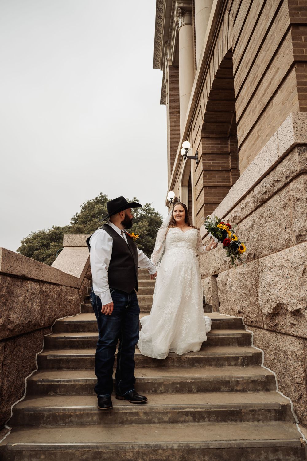 A couple in western wedding attire poses on grand stone steps of a historic building.