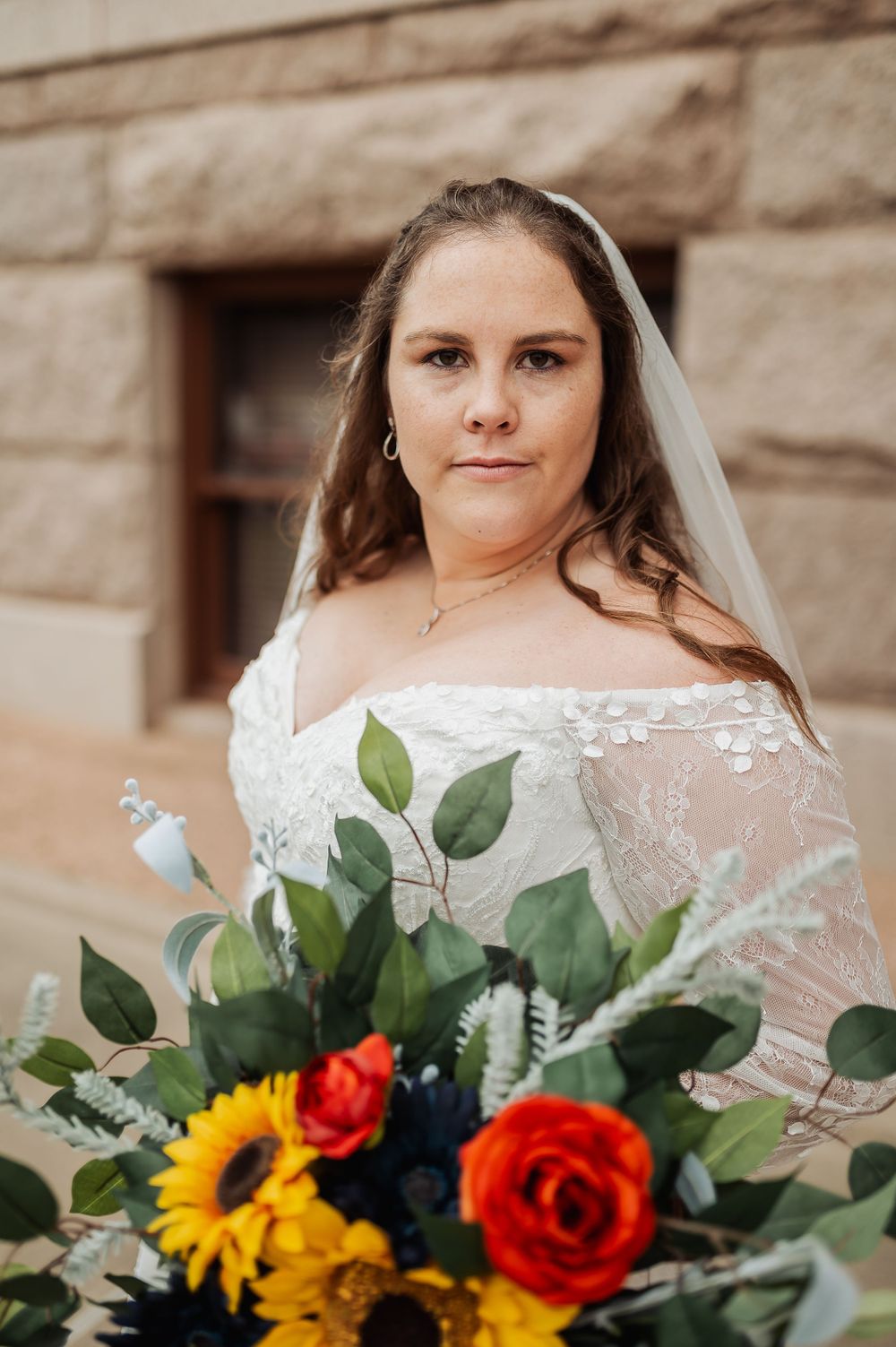 A bride in a lace off-shoulder wedding dress holds a vibrant bouquet with sunflowers and roses against a stone wall.