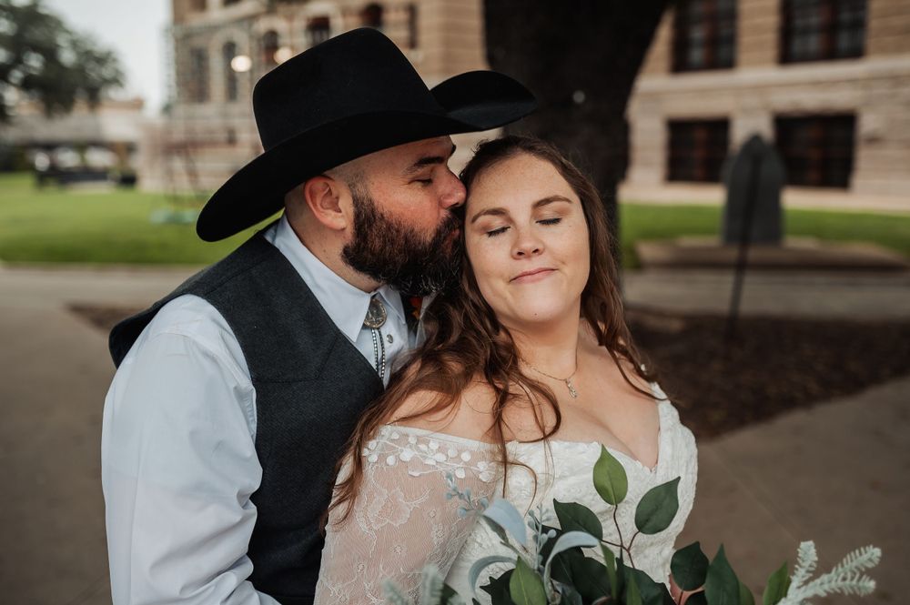 A western-styled wedding couple share an intimate moment outdoors with the groom in a cowboy hat.