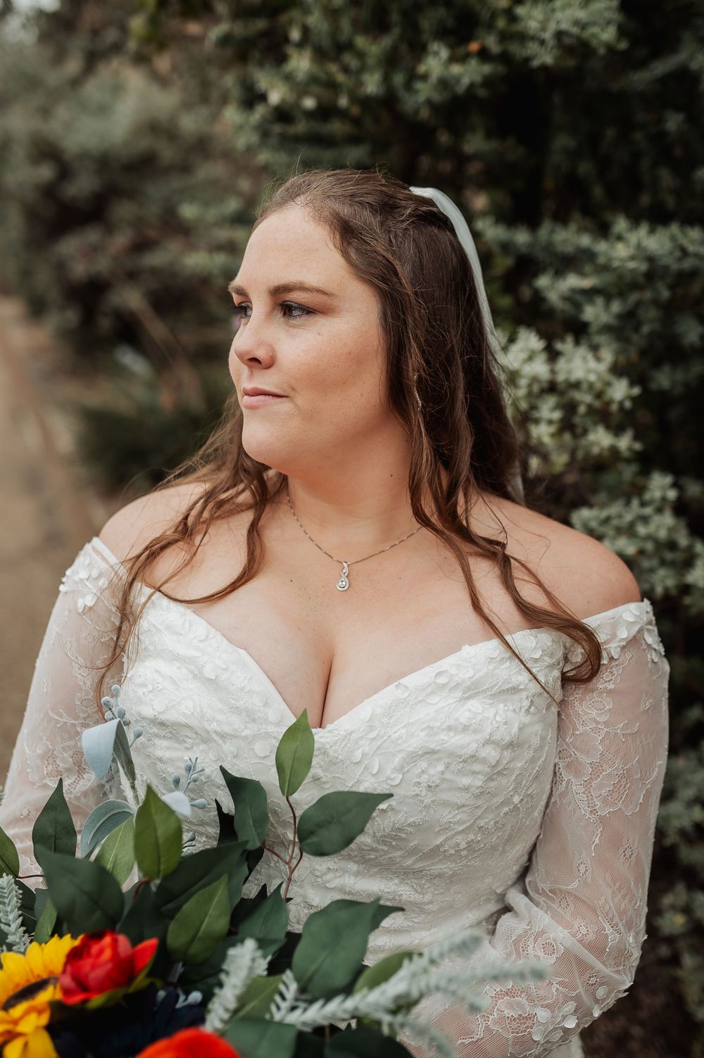 A bride wearing an off-shoulder lace gown poses with a colorful bouquet featuring sunflowers and roses.