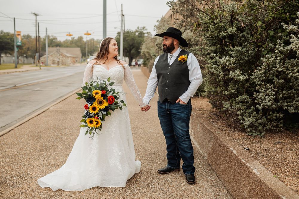 A bride in white lace and groom in western attire walk together on a path holding a bright floral bouquet.