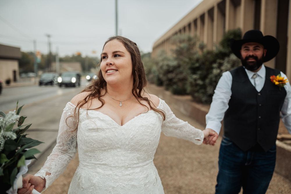 A joyful bride in lace walks outdoors while her western-styled groom follows behind.