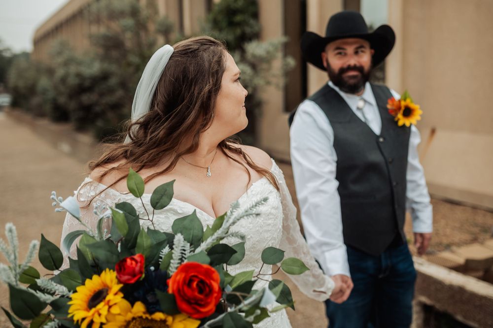 A romantic wedding photo features western-styled couple with sunflower bouquet against brick building.