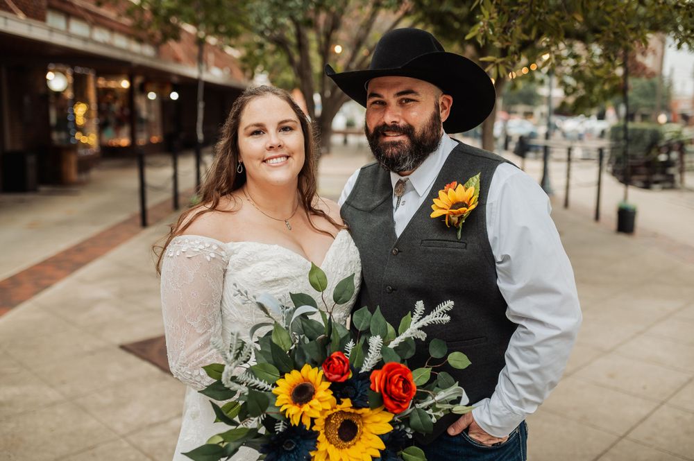 A happy wedding couple in western attire pose with a colorful bouquet of sunflowers and roses on a city street.