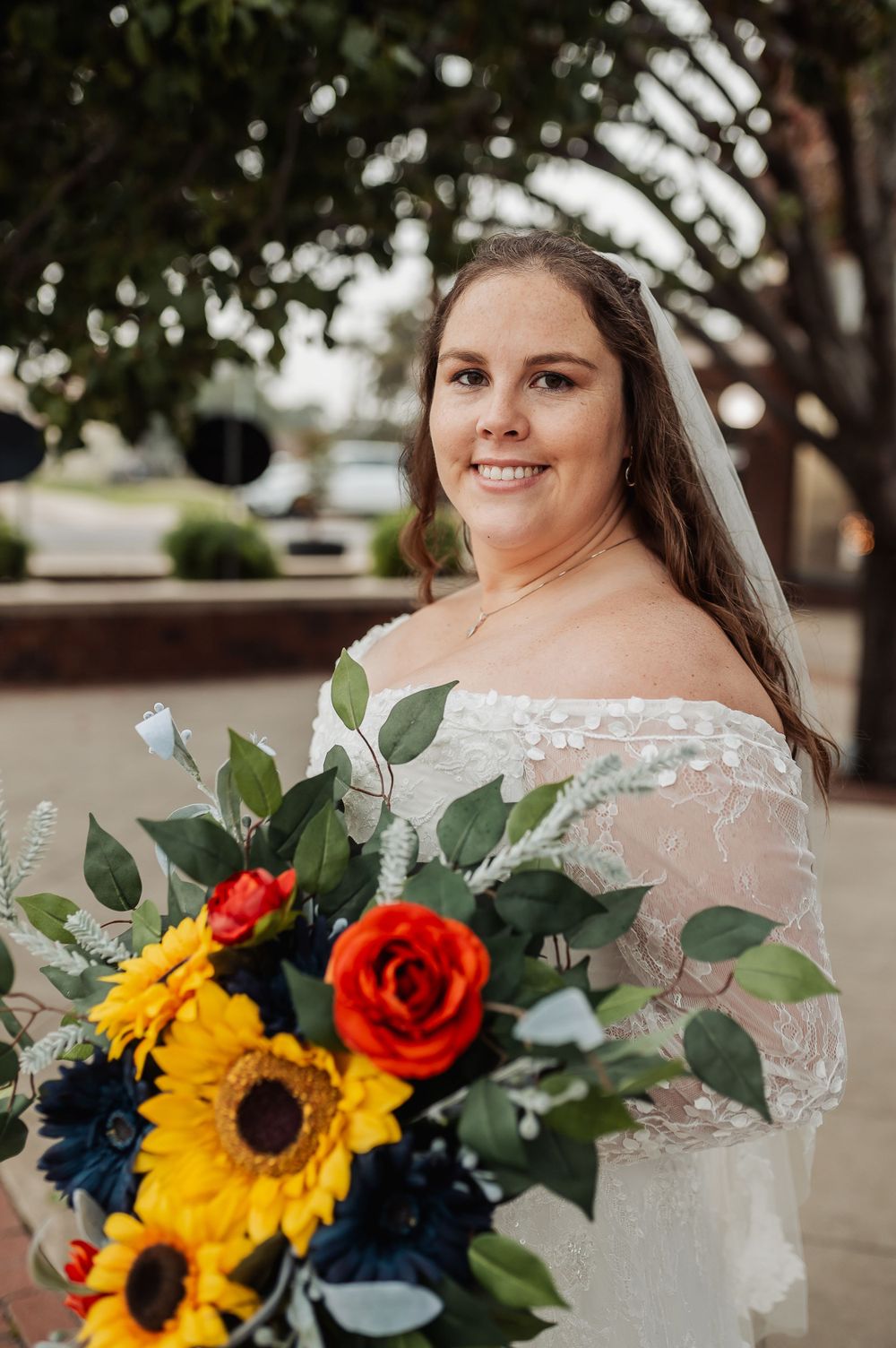 A radiant bride holds a vibrant bouquet of sunflowers and roses while wearing an off-shoulder lace gown.