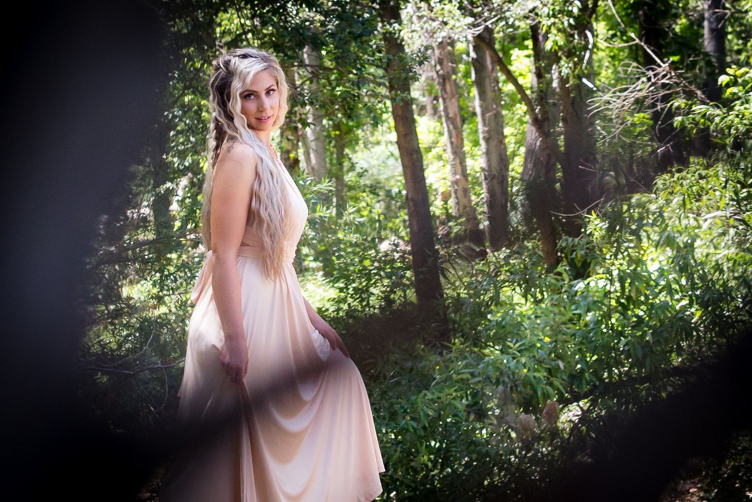 A dreamy portrait in a sun-dappled forest shows someone in an ethereal blush pink dress among the trees.