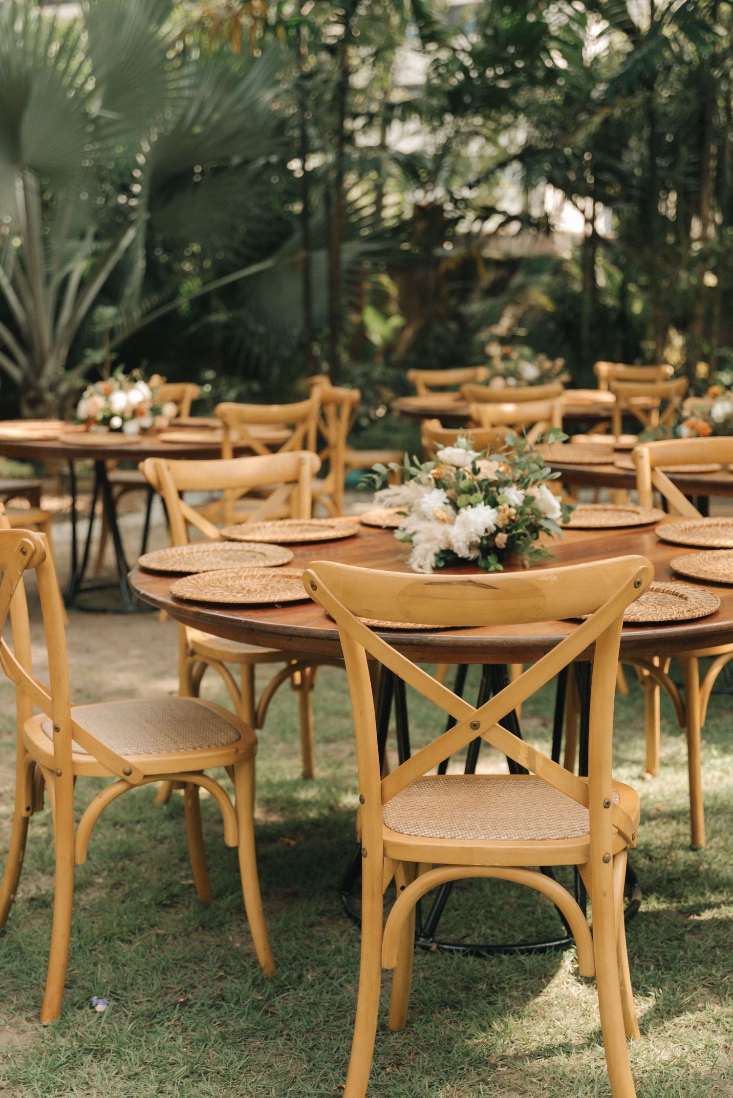 Outdoor wedding reception setup with wooden cross-back chairs and floral centerpieces.