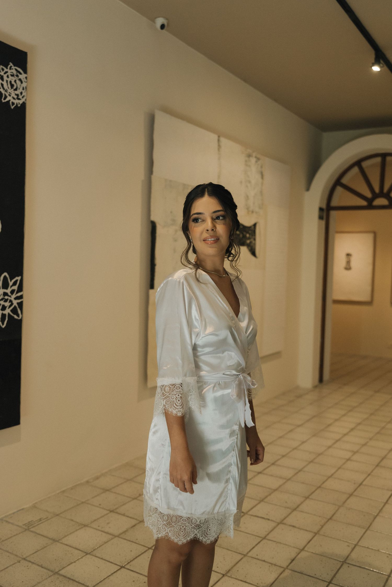 Person in white silk robe with lace trim standing in a modern gallery hallway.