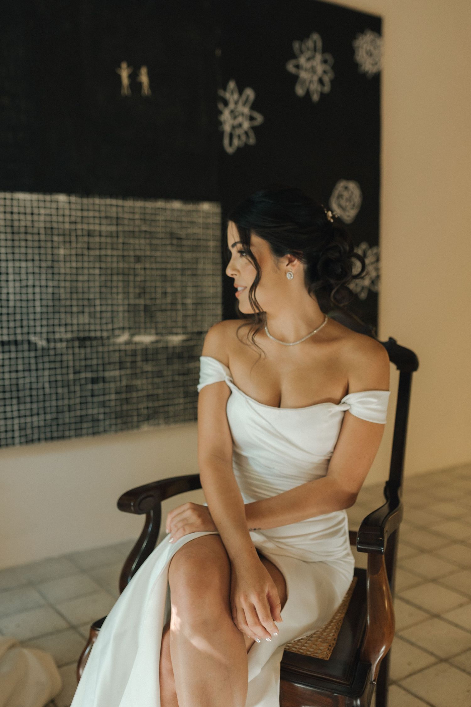 Person in off-shoulder white dress seated in wooden chair against textured wall.