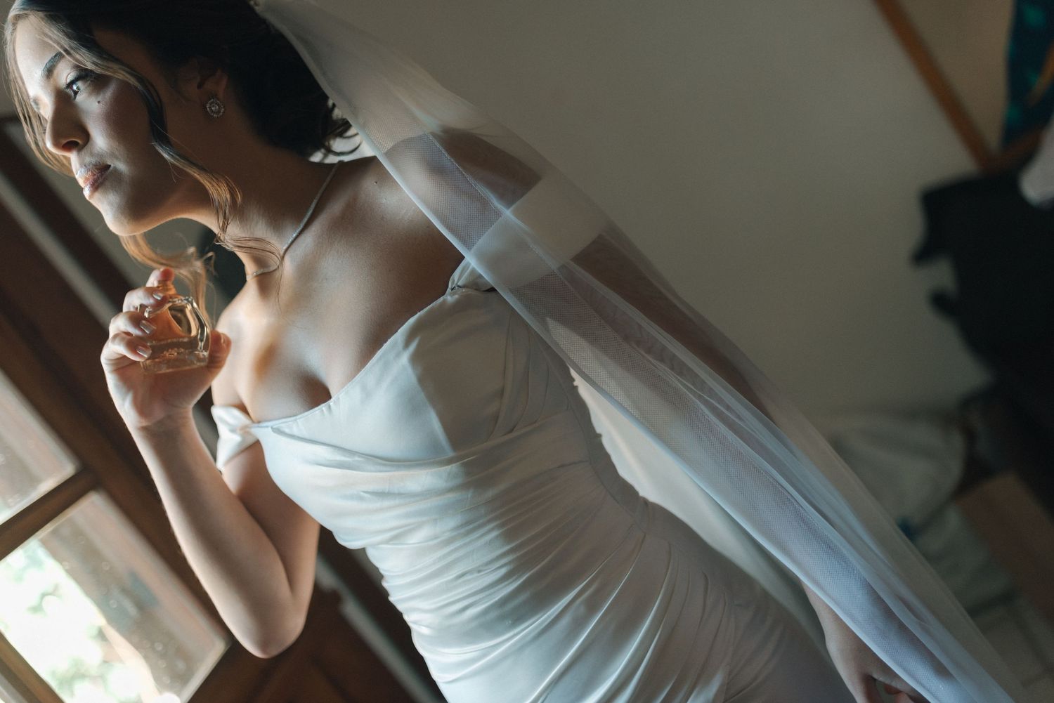 Person in fitted white dress adjusting veil in soft window light.