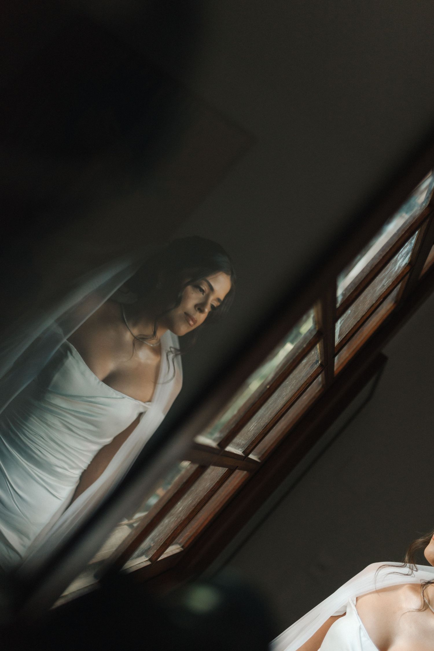 A moody bridal portrait showing a woman in a white dress gazing downward near a window.